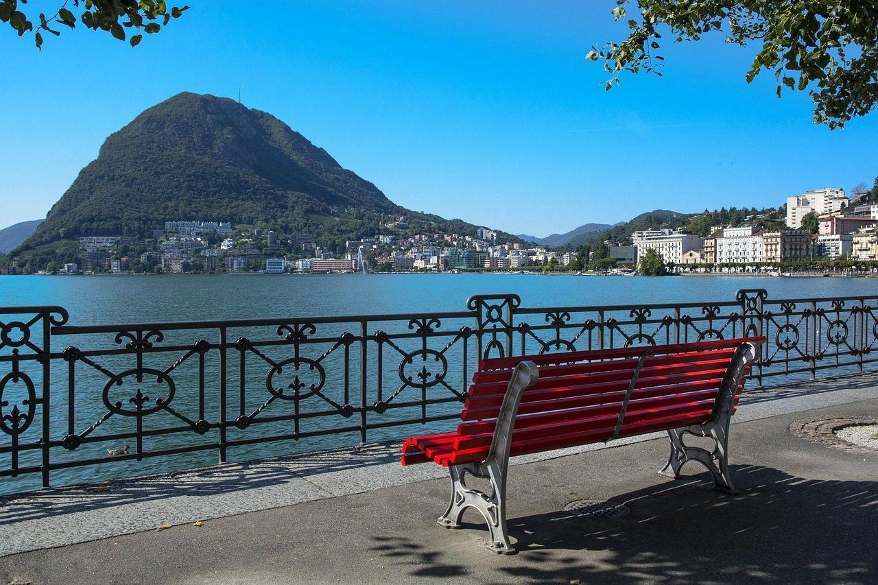 Paseo junto al lago de Lugano con un banco rojo y vista al Monte San Salvatore